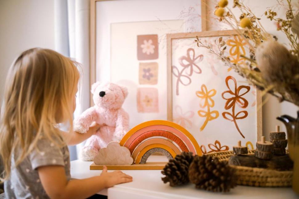 A little girl with blonde hair stands at a low shelf with artwork and a small teddy bear.